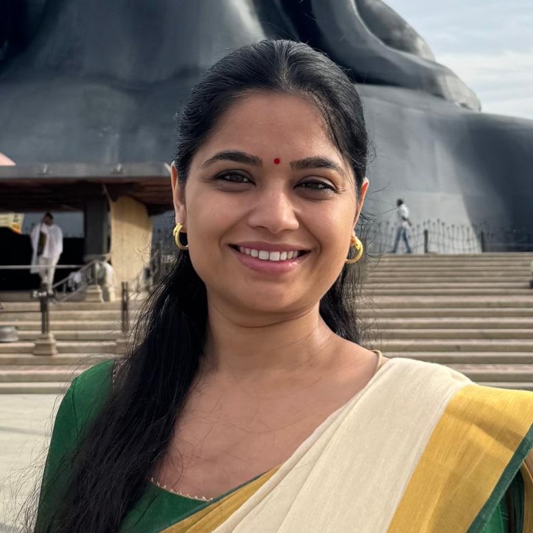 A woman smiling while wearing a traditional sari, standing in front of a large statue. The background features steps leading to the statue and people in the distance.