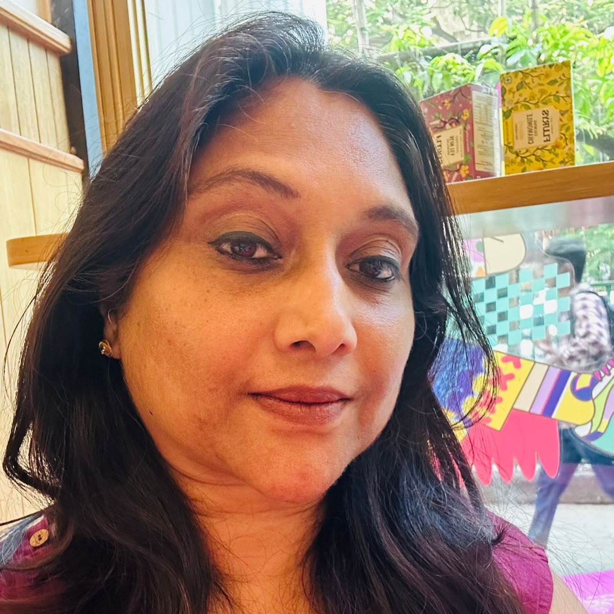 A close-up portrait of a woman with long black hair, wearing a pink top, smiling softly. There are colorful boxes visible in the background along with some greenery.